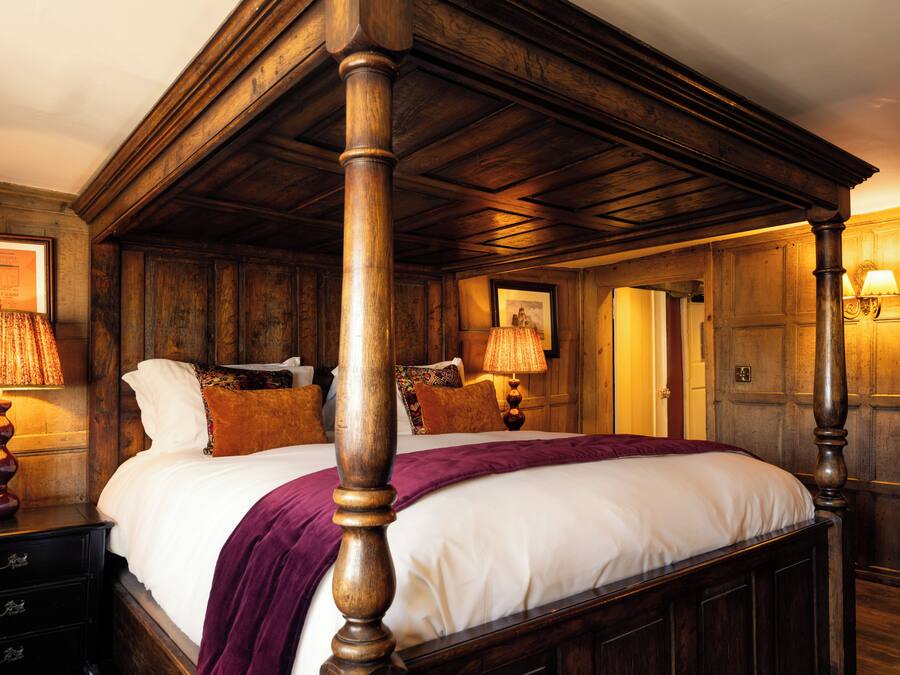A traditional four-poster bed with a wooden headboard, a white bedspread, and a purple throw blanket. There are two bedside lamps with lampshades, a framed picture on the wall, and a wooden dresser with drawers.