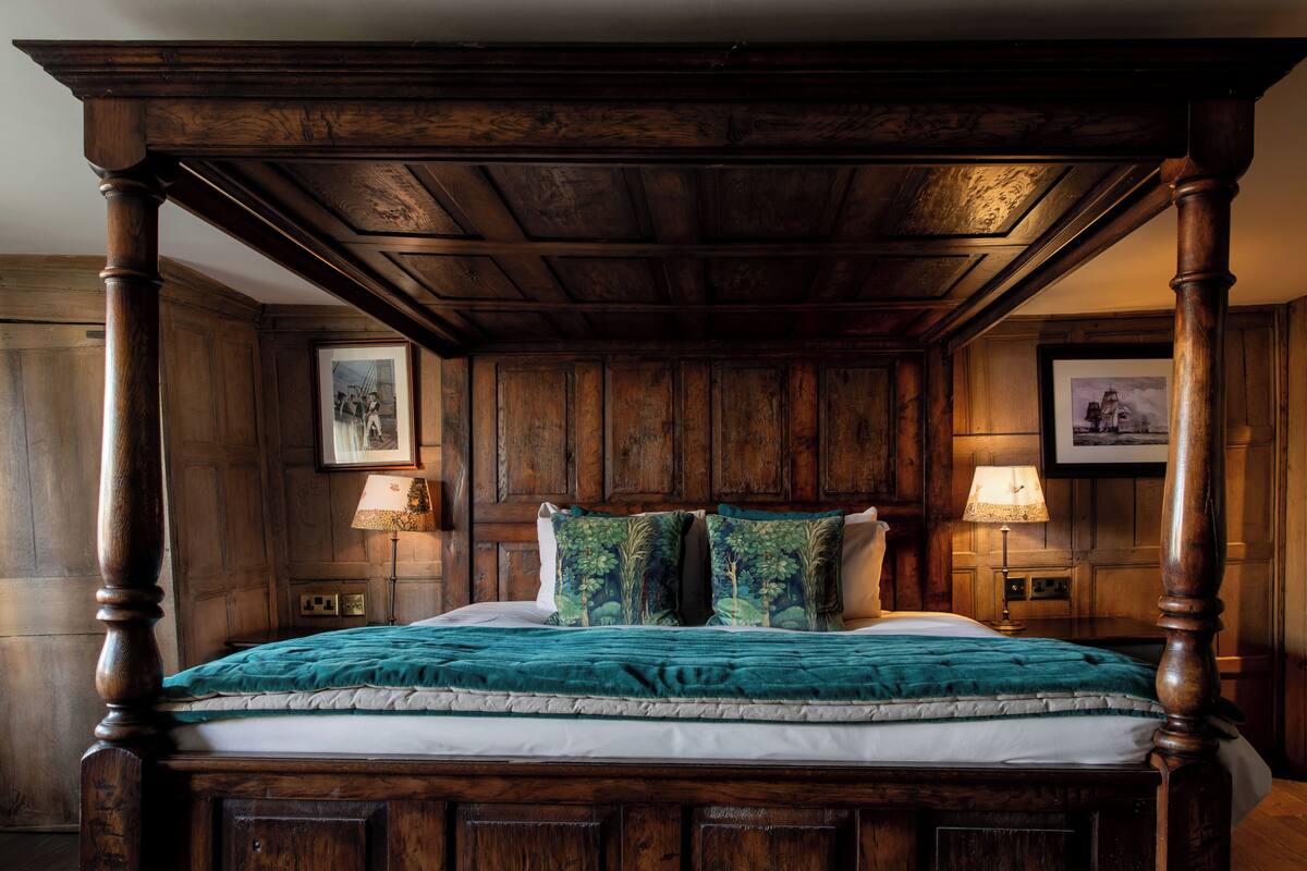 A traditional wooden four-poster bed with a teal bedspread and matching pillows, flanked by two bedside tables with lamps.