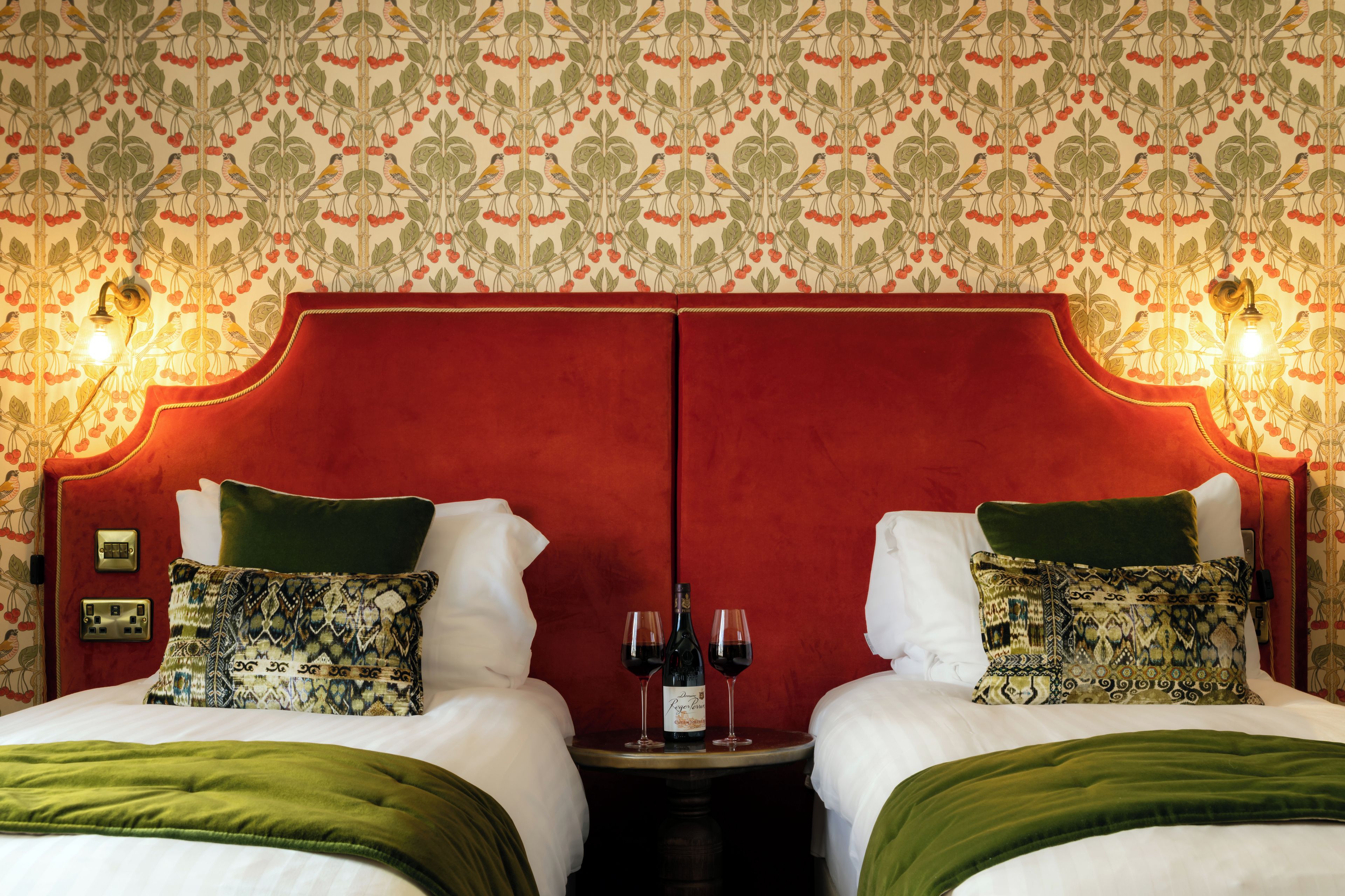 A hotel room with a mirrored headboard, two beds with green and patterned pillows, a bedside table with wine glasses, and a wall with a repeating bird and fruit pattern.