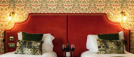A hotel room with a mirrored headboard, two beds with green and patterned pillows, a bedside table with wine glasses, and a wall with a repeating bird and fruit pattern.
