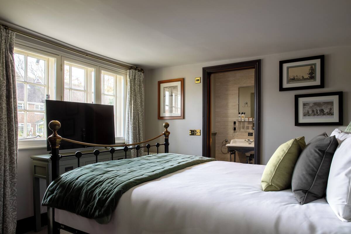 A neatly made bed with a green blanket and pillows, a window with curtains, and framed pictures on the wall.