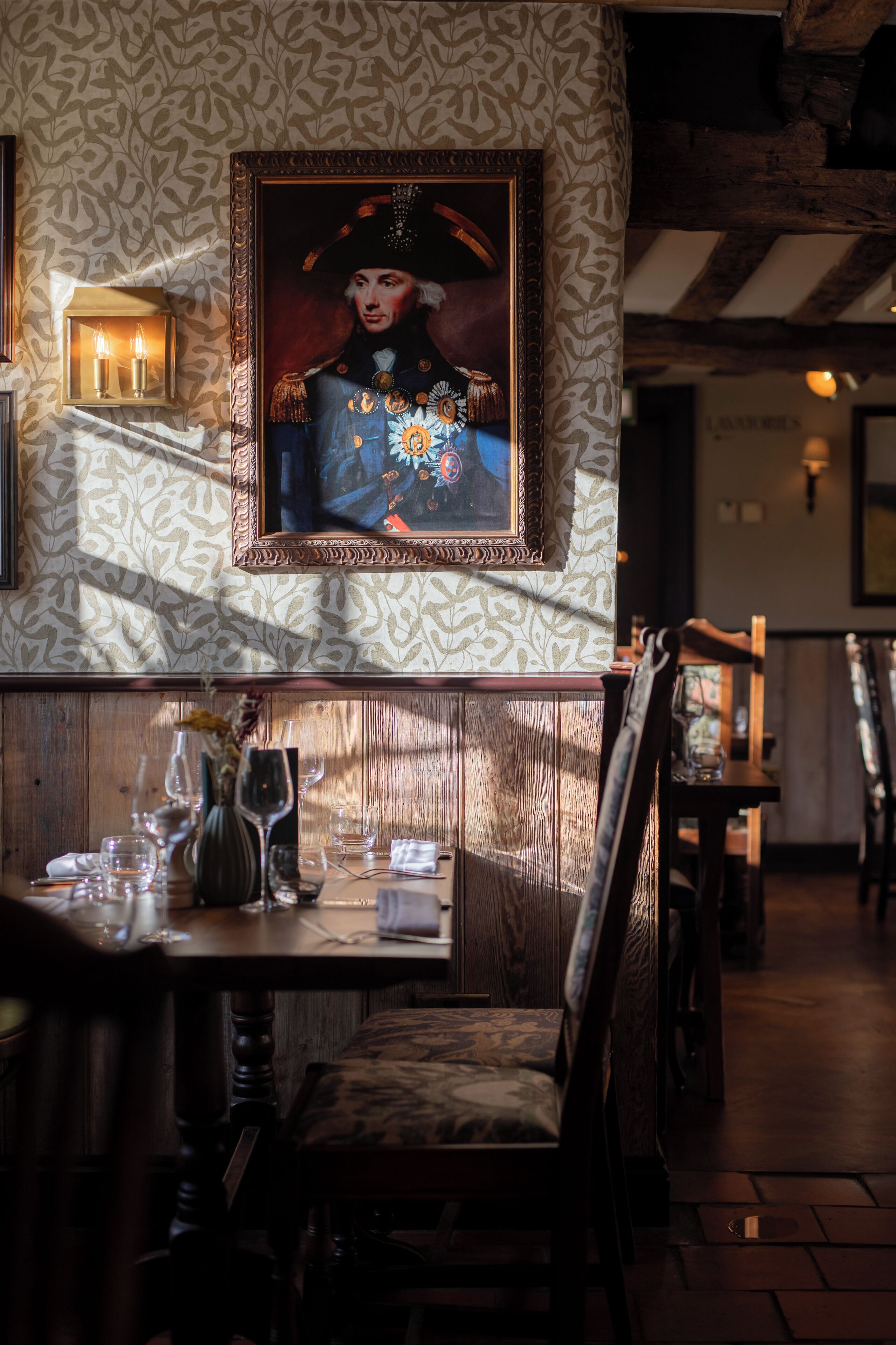 Een restaurantinterieur met een houten bank, een gedekte tafel met glazen en een vaas, en een lijst met een portret aan de muur.