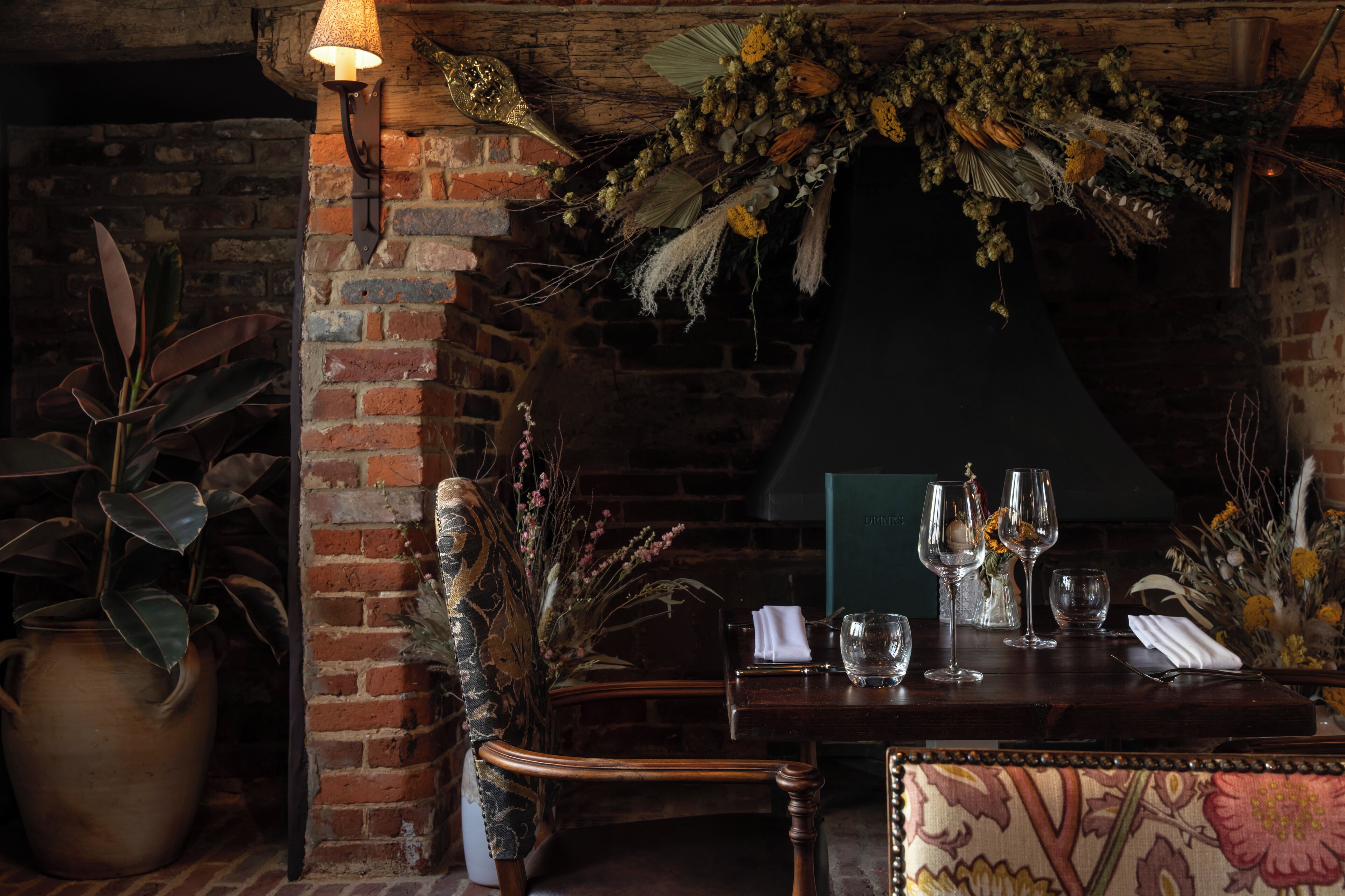 A rustic dining area with a brick wall, wooden furniture, and a table set with wine glasses and a vase of flowers.