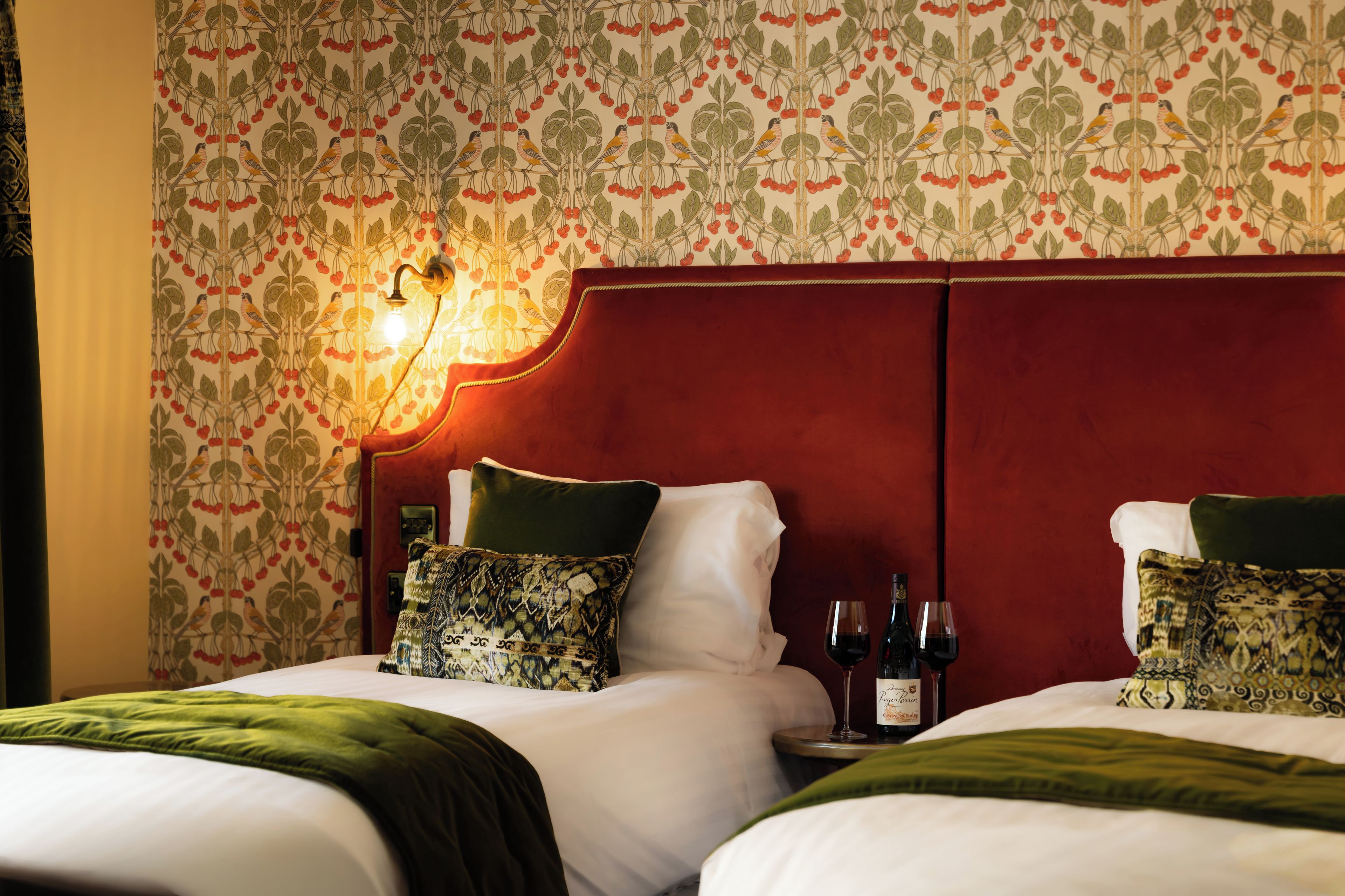 A hotel room with two beds, a red headboard, patterned wallpaper, and a nightstand with a wine bottle and glass.