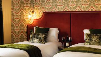 A hotel room with two beds, a red headboard, patterned wallpaper, and a nightstand with a wine bottle and glass.
