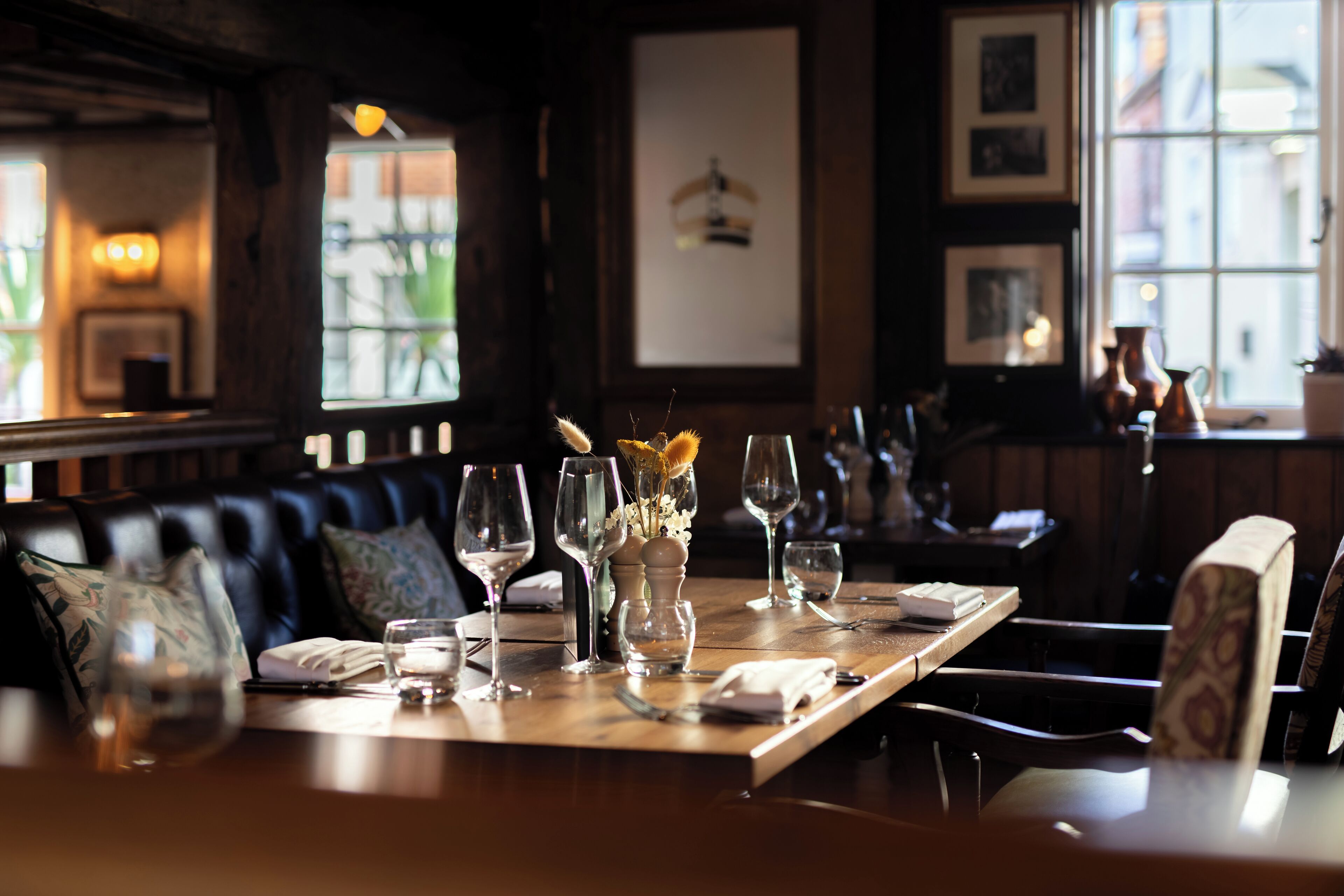 A restaurant setting with a wooden table set for dining, featuring wine glasses, a vase with flowers, and a menu.