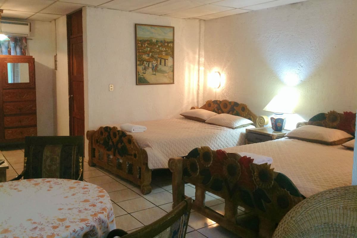 A hotel room with two beds, a wooden headboard, a painting on the wall, and a table with a floral tablecloth.