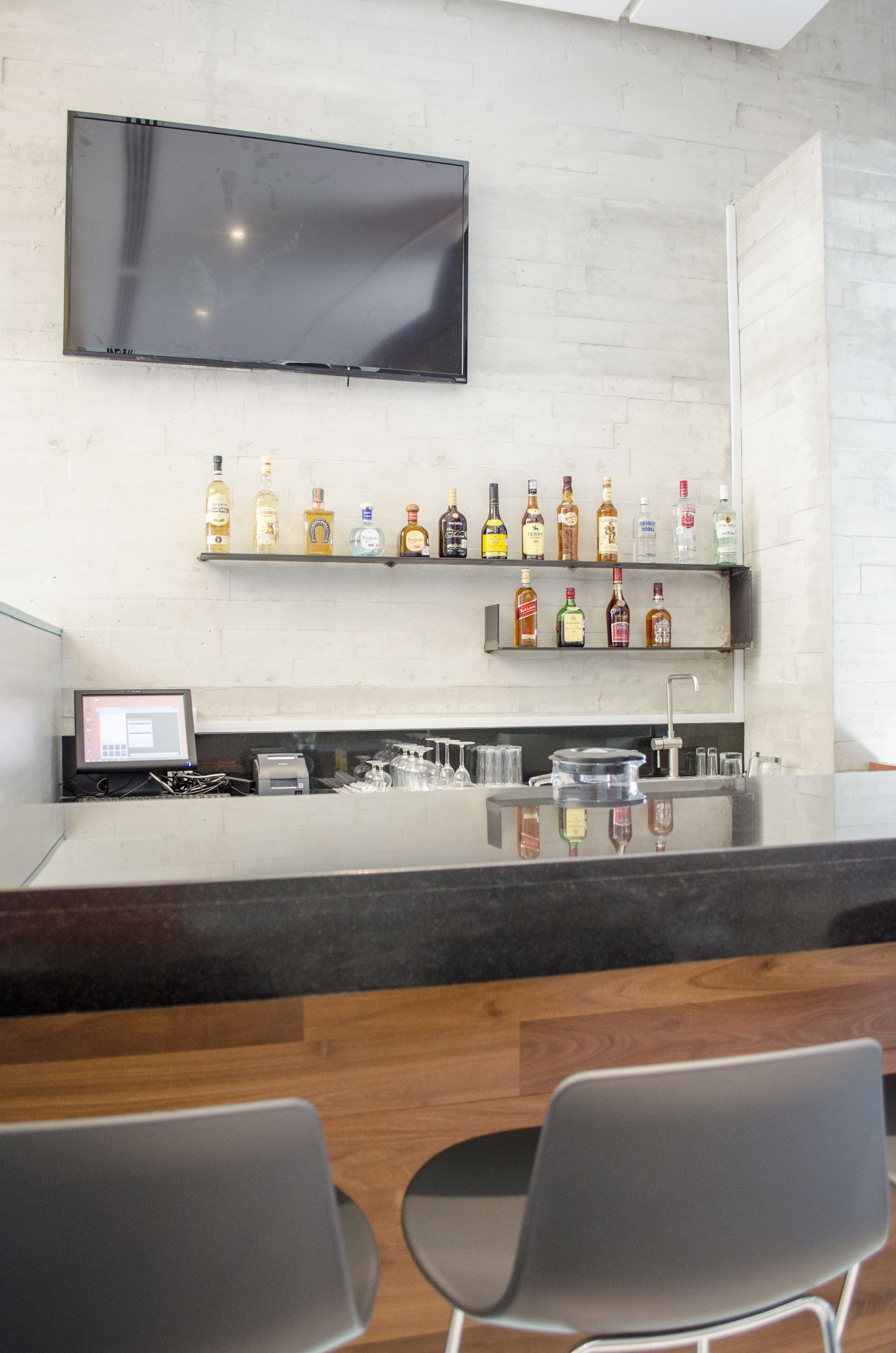A bar area with a flat-screen TV mounted on the wall, shelves with various liquor bottles, a cash register, and two chairs.