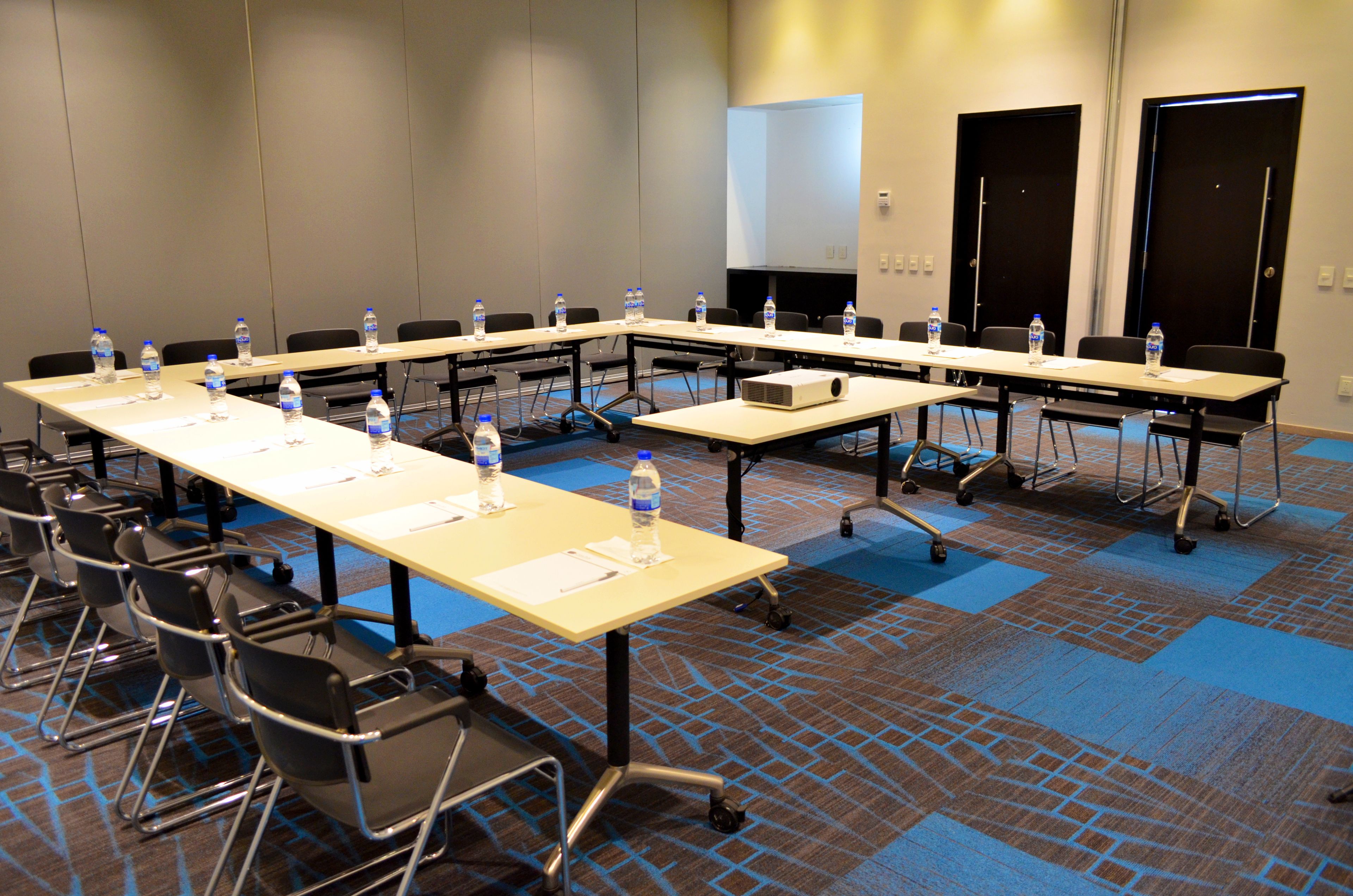Sala de conferência com mesas longas, cadeiras, garrafas de água e um projetor.