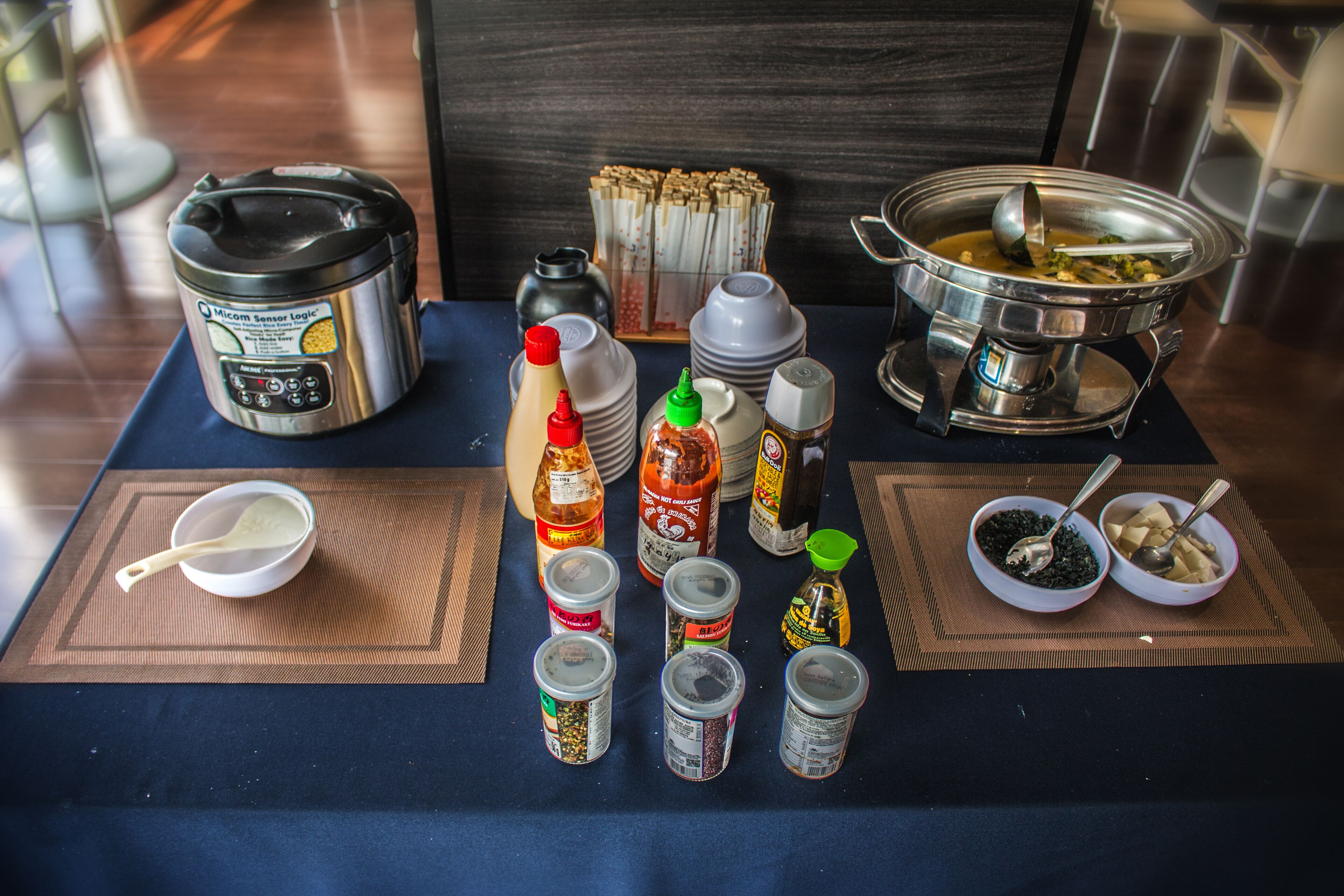 A table with various condiments, a rice cooker, and a soup pot, suggesting a self-service dining setup.