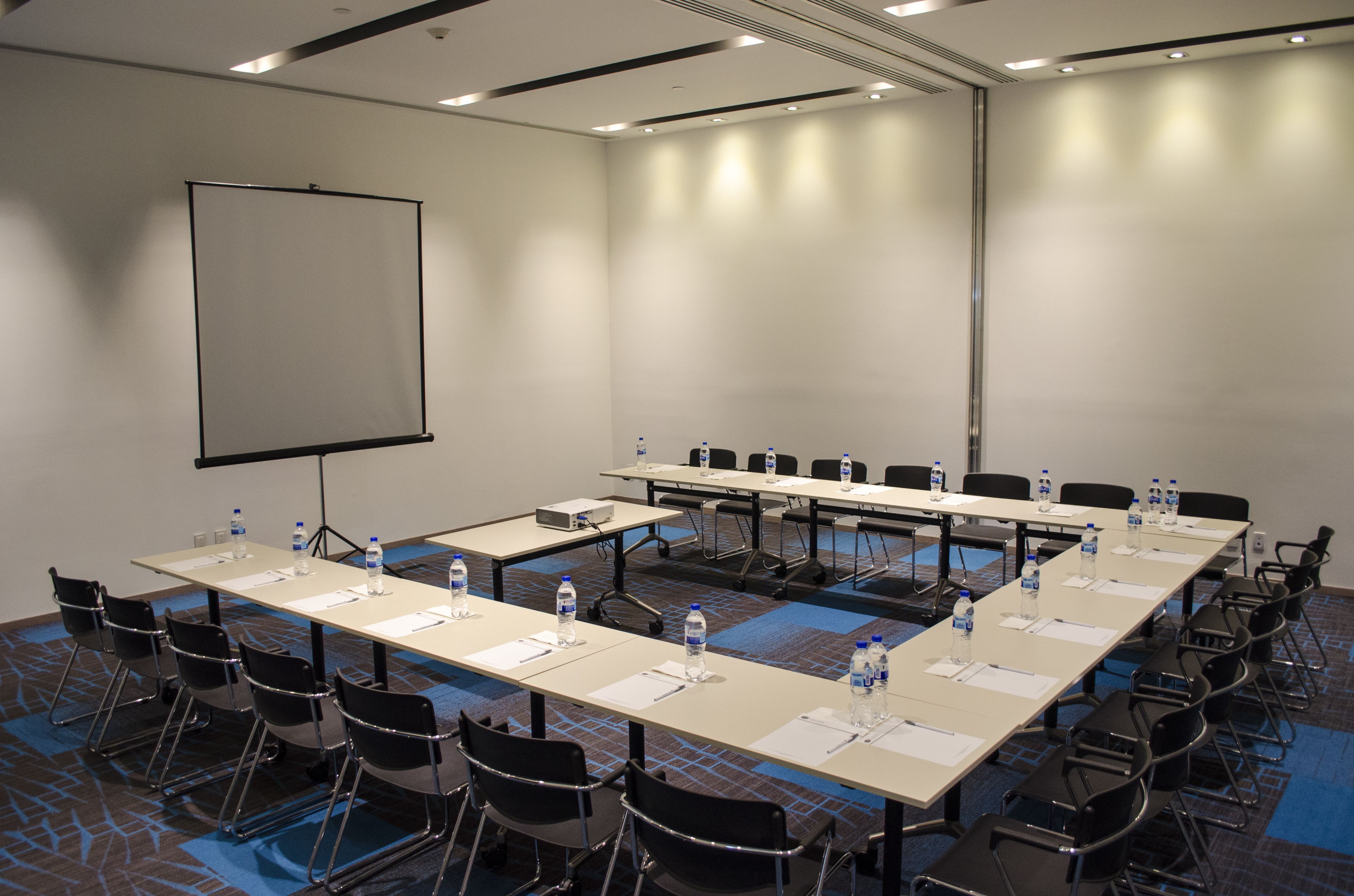 Una sala conferenze con un grande schermo, diversi tavoli con sedie, bottiglie d'acqua e un pavimento rivestito di moquette.