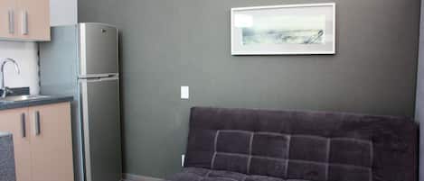 Una kitchenette compacta con refrigerador y lavamanos, un sofá de color oscuro y un cuadro en la pared.