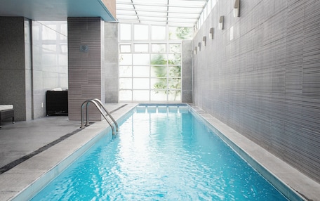 An indoor swimming pool with clear glass walls, a metal ladder, and a bench.