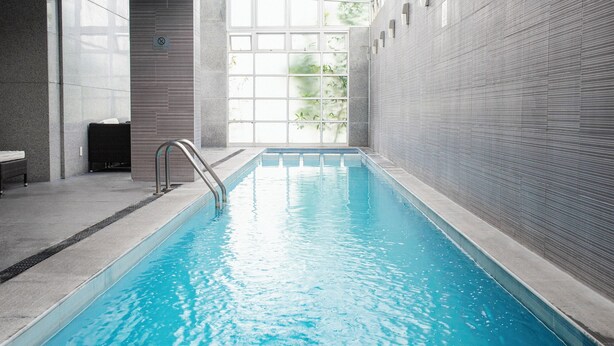 An indoor swimming pool with clear glass walls, a metal ladder, and a bench. - Hotel Casa Inn Galerias (Celaya)