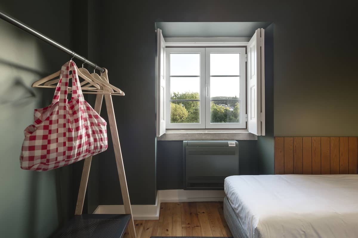 A bedroom with a bed, a window, a radiator, a wooden floor, a grey mat, and a red and white checkered bag hanging on a wooden hanger.