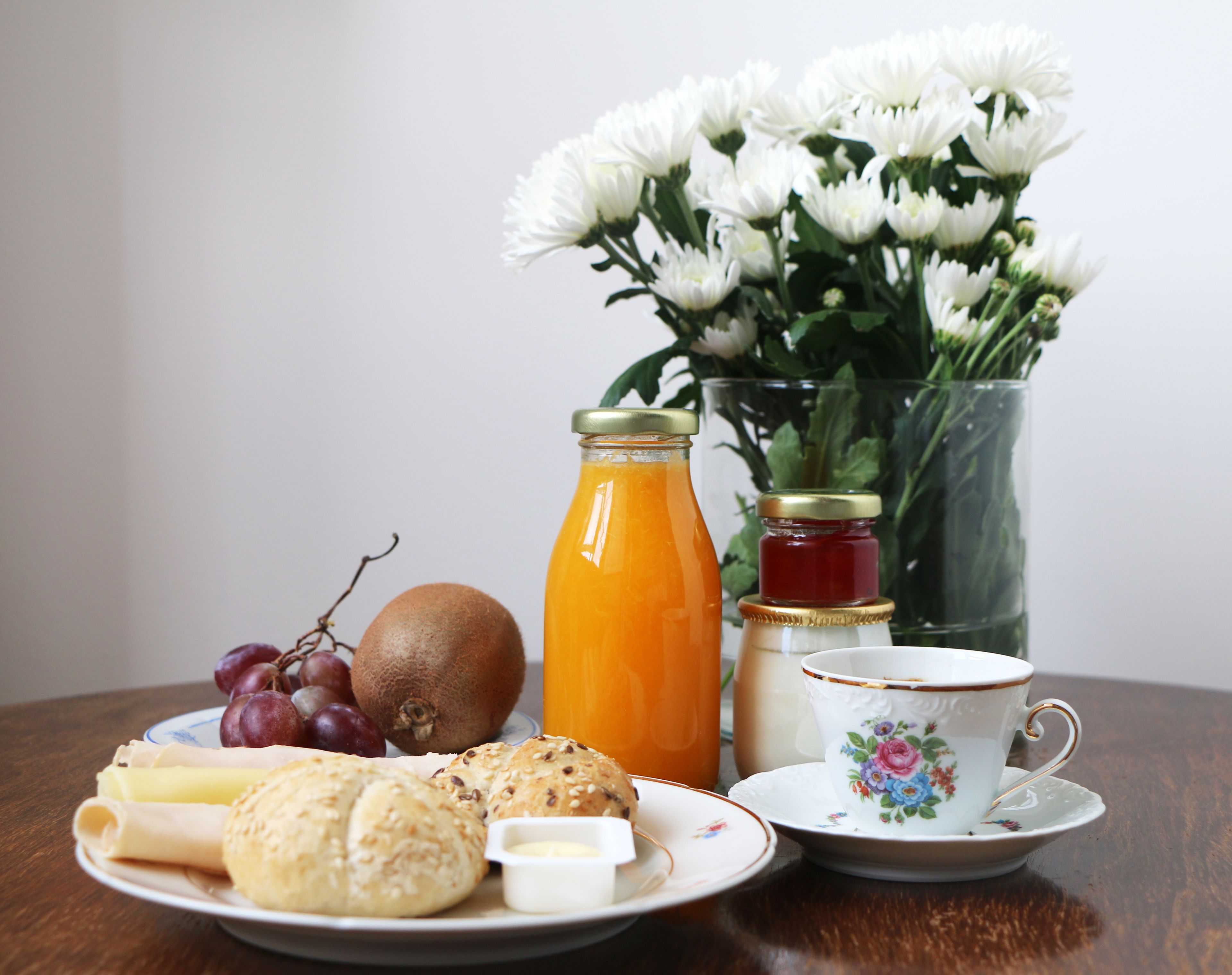 A table with a plate of food, a glass of orange juice, a jar of jam, a cup of tea, and a vase of flowers.