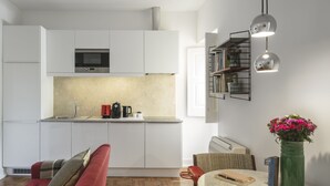 Une cuisine moderne avec des meubles blancs, un four à micro-ondes et une machine à café. On y trouve un coin repas avec une table ronde, des chaises et un canapé rouge. Un vase vert rempli de fleurs est posé sur la table.