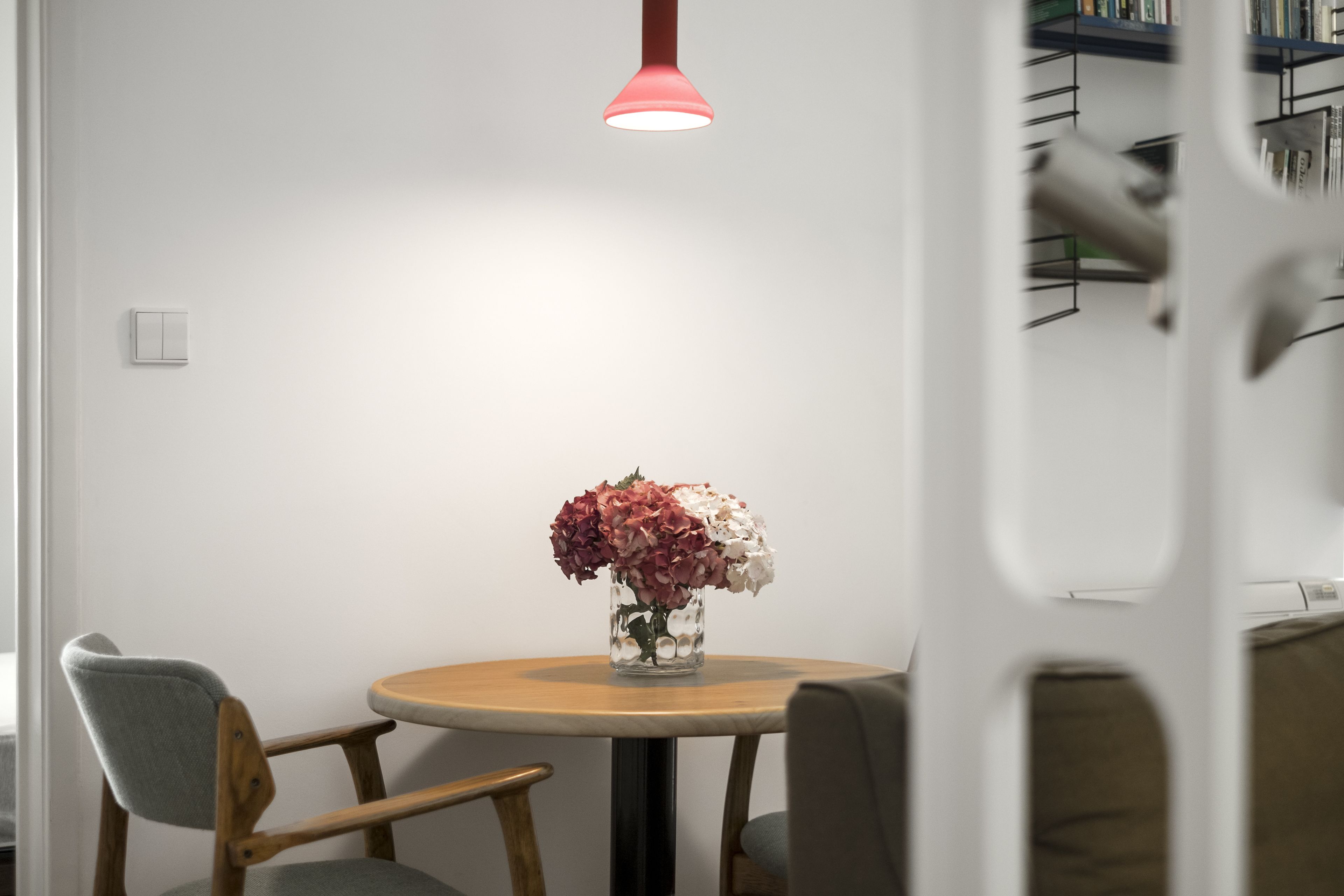 Une table ronde en bois, sur laquelle est posé un vase transparent rempli de fleurs, une chaise en bois et une suspension moderne.