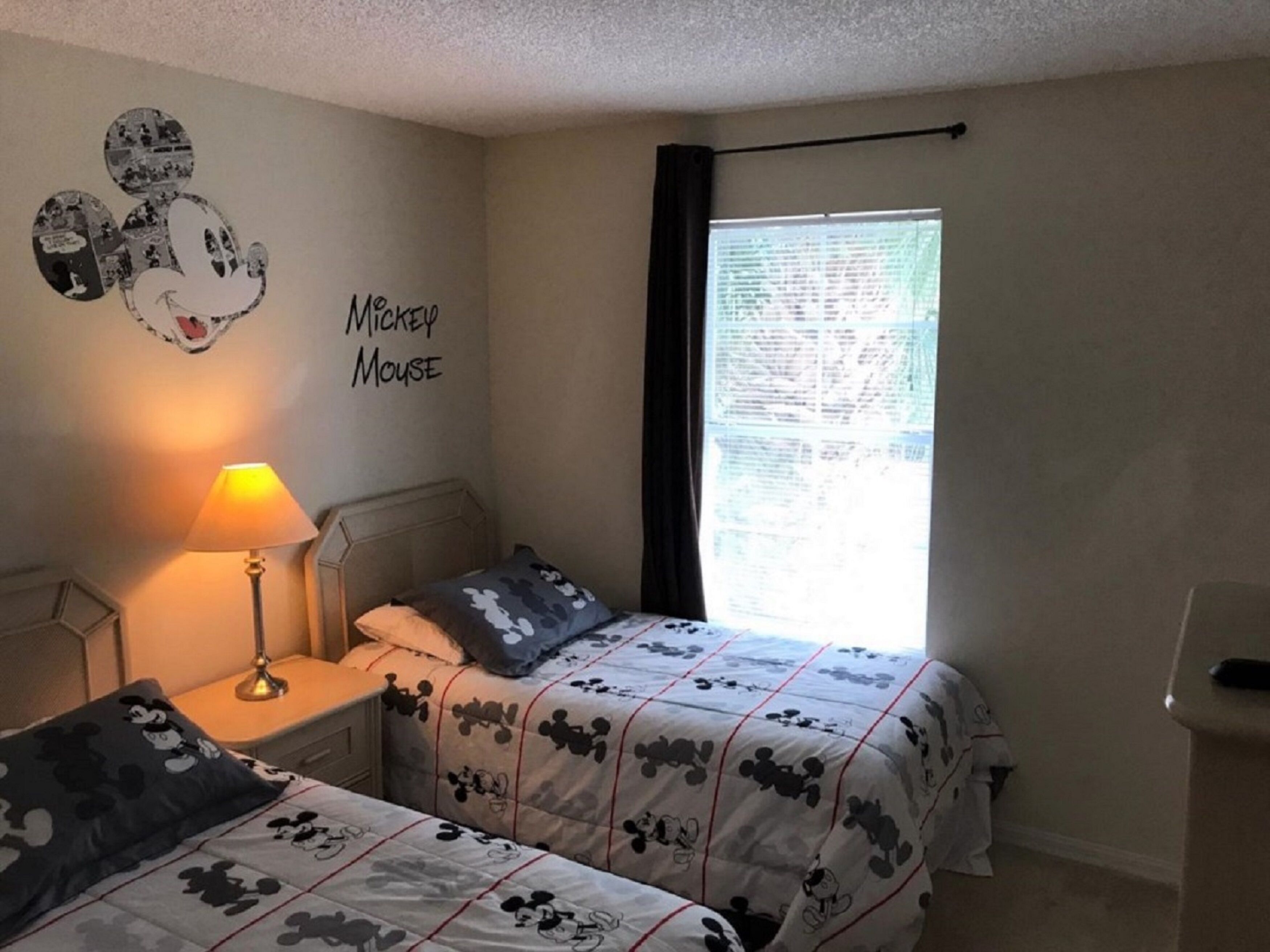 A bedroom with two beds, a nightstand, a lamp, a Mickey Mouse wall decal, and a window with curtains.