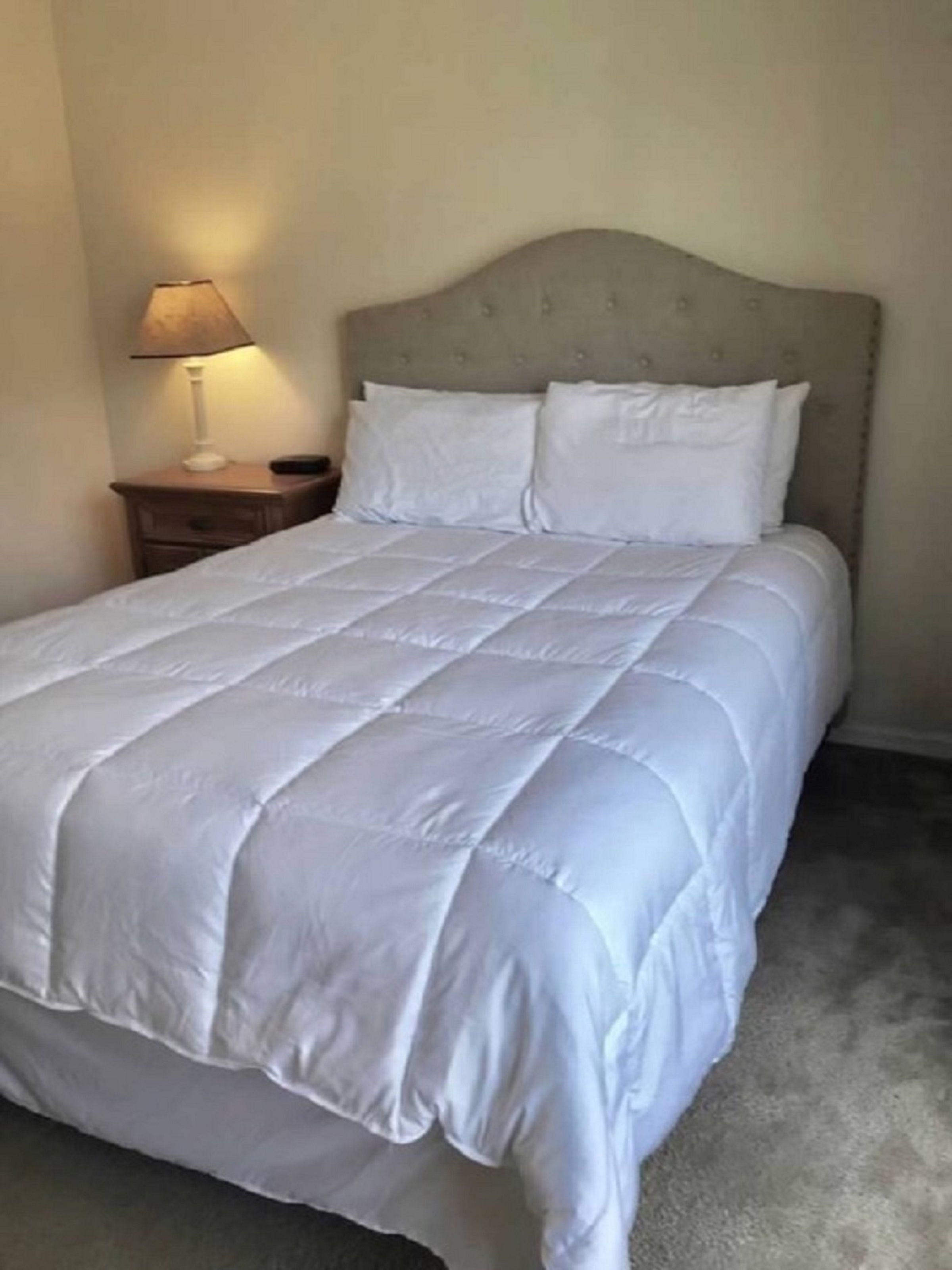 Una cama bien hecha con edredón blanco acolchado, una mesita de noche de madera con una lámpara y un cabecero con diseño capitonado.