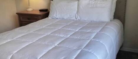 Una cama bien hecha con edredón blanco acolchado, una mesita de noche de madera con una lámpara y un cabecero con diseño capitonado.