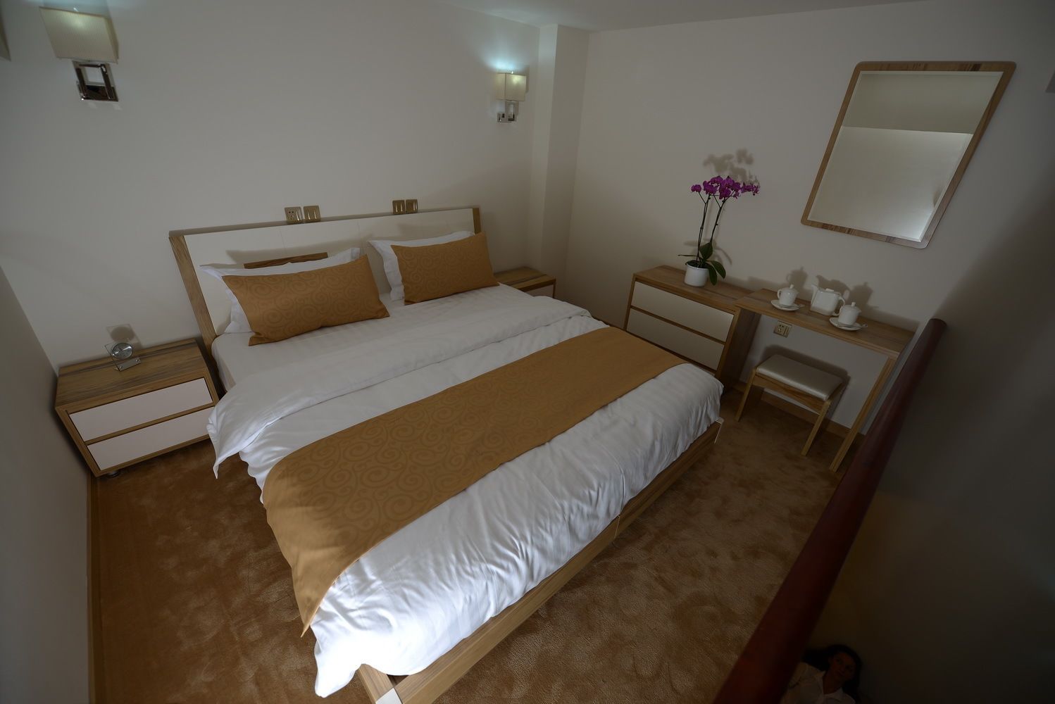 A hotel room with a large bed, a bedside table with a lamp, a small desk with a chair, and a mirror on the wall.