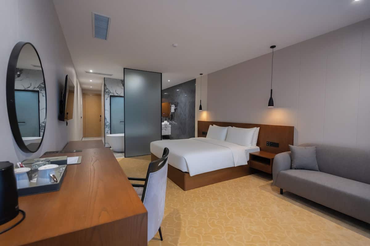 A modern hotel room with a large bed, a sofa, a desk with a mirror, and a television.