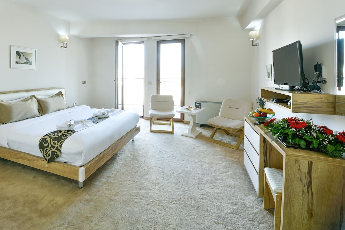A hotel room with a bed, bedside tables, a desk with a chair, a television, and a large window.