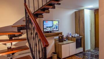 A wooden staircase with a black railing leading to a living area with a television, a kitchen area, and a wooden wardrobe.