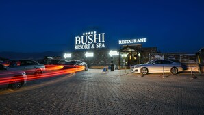 A nighttime view of the BUSHI RESORT & SPA entrance with a restaurant sign, cars parked in front, and a paved driveway. - Bushi Resort & Spa (Skopje)