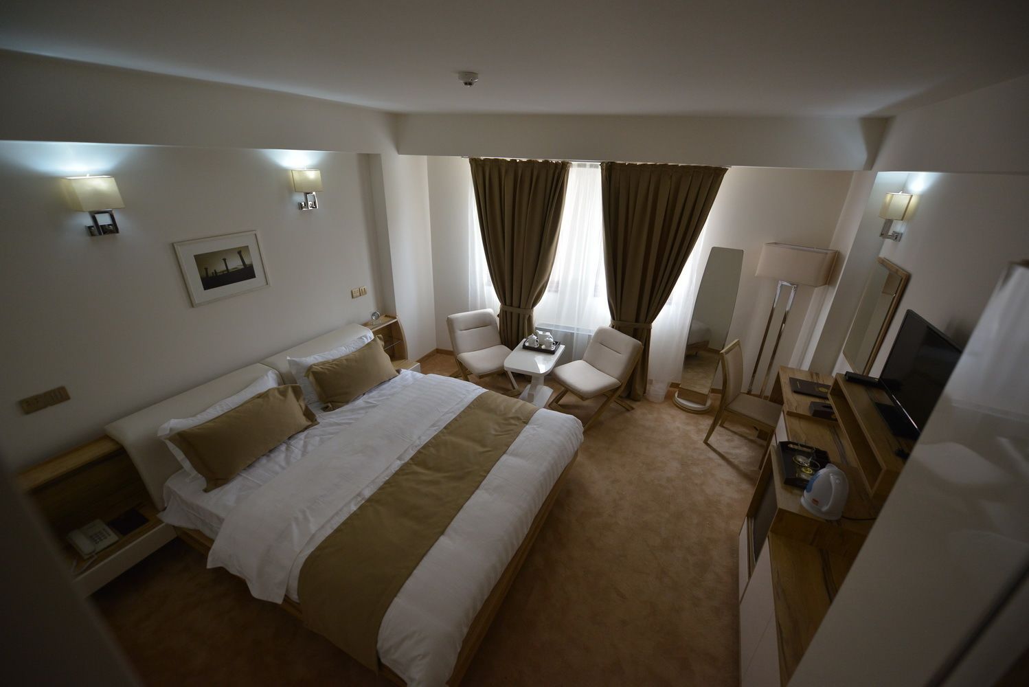 A hotel room with a large bed, two chairs, a small table, a TV, and a desk with a lamp.