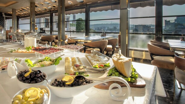 A buffet setup with various food items and a view of the city through large windows. - Bushi Resort & Spa (Skopje)