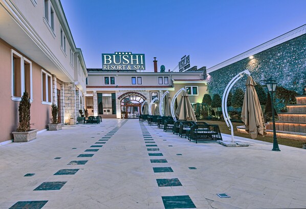 A hotel named BUSHI RESORT & SPA with a central entrance, outdoor seating area with umbrellas, and a tiled walkway. - Bushi Resort & Spa (Skopje)