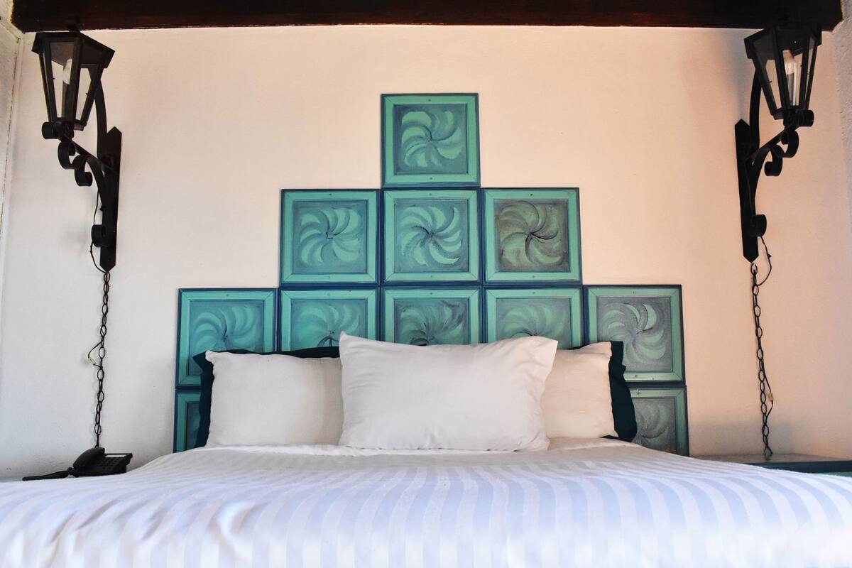 A bed with white bedding, a patterned blue and white floor, and a wall with teal square art pieces.