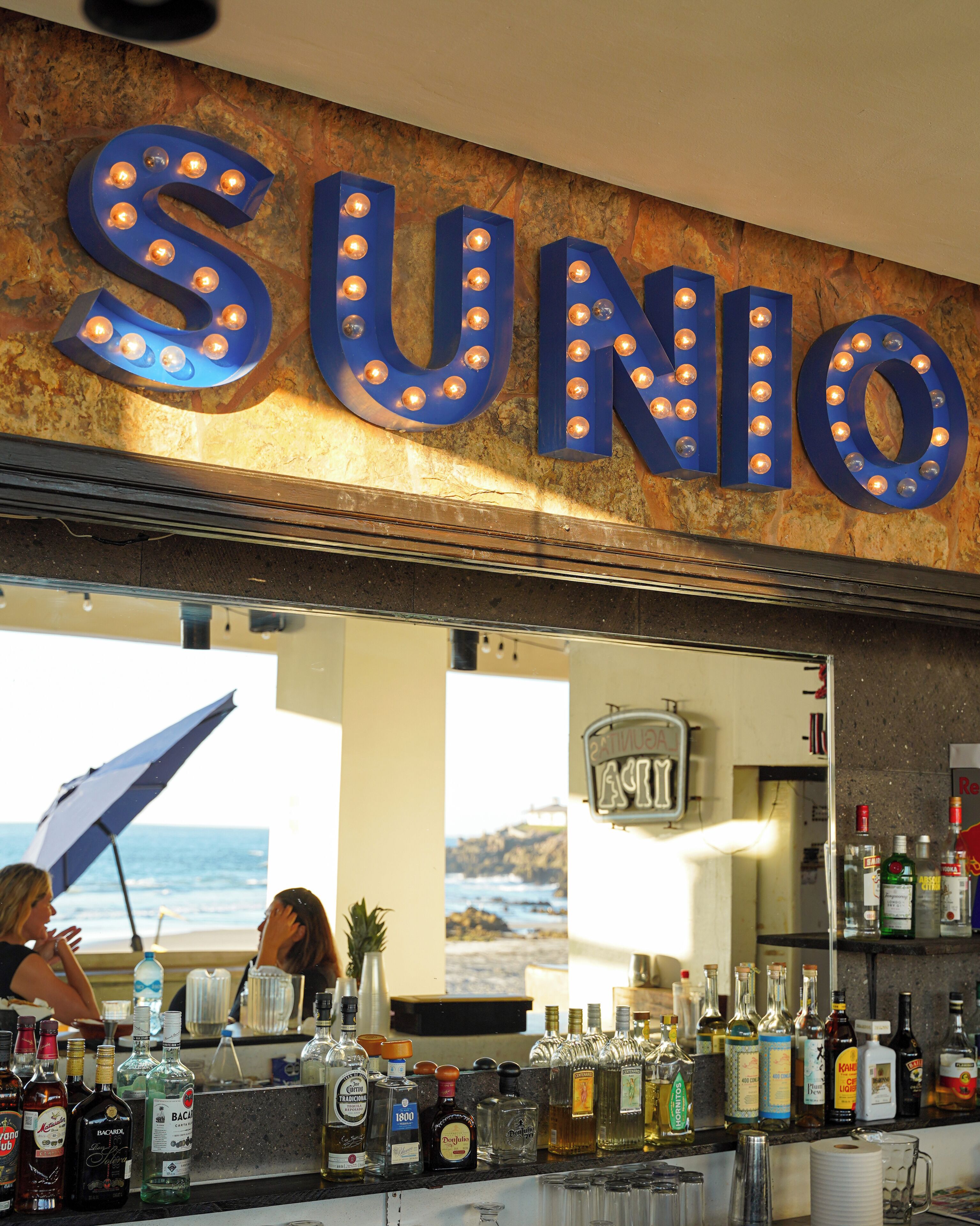 A bar with a neon sign reading 'SUNIO' and a view of the ocean through the window.