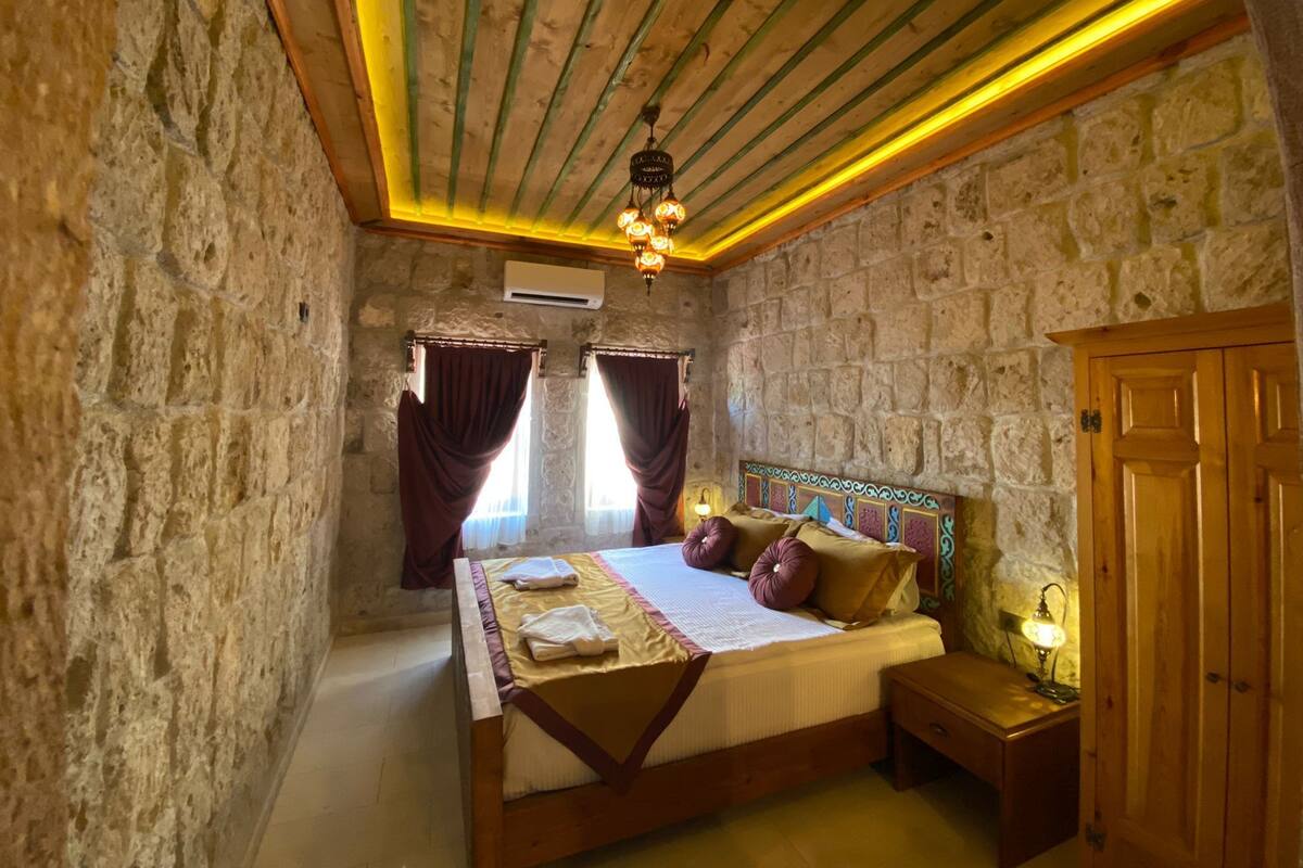A bedroom with a stone wall, wooden ceiling beams, a bed with pillows, a nightstand, and a lamp.
