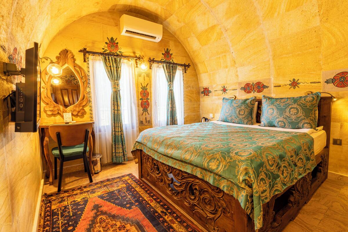 A bedroom with a wooden bed, a desk with a chair, a large mirror, and a patterned rug.