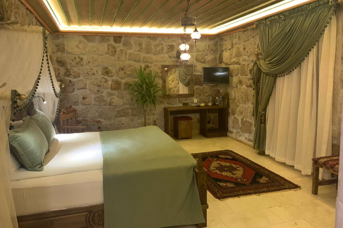 A bedroom with a stone wall, a bed with a green bedspread, a hanging chair, a desk with a mirror, a television, and a red and brown rug.