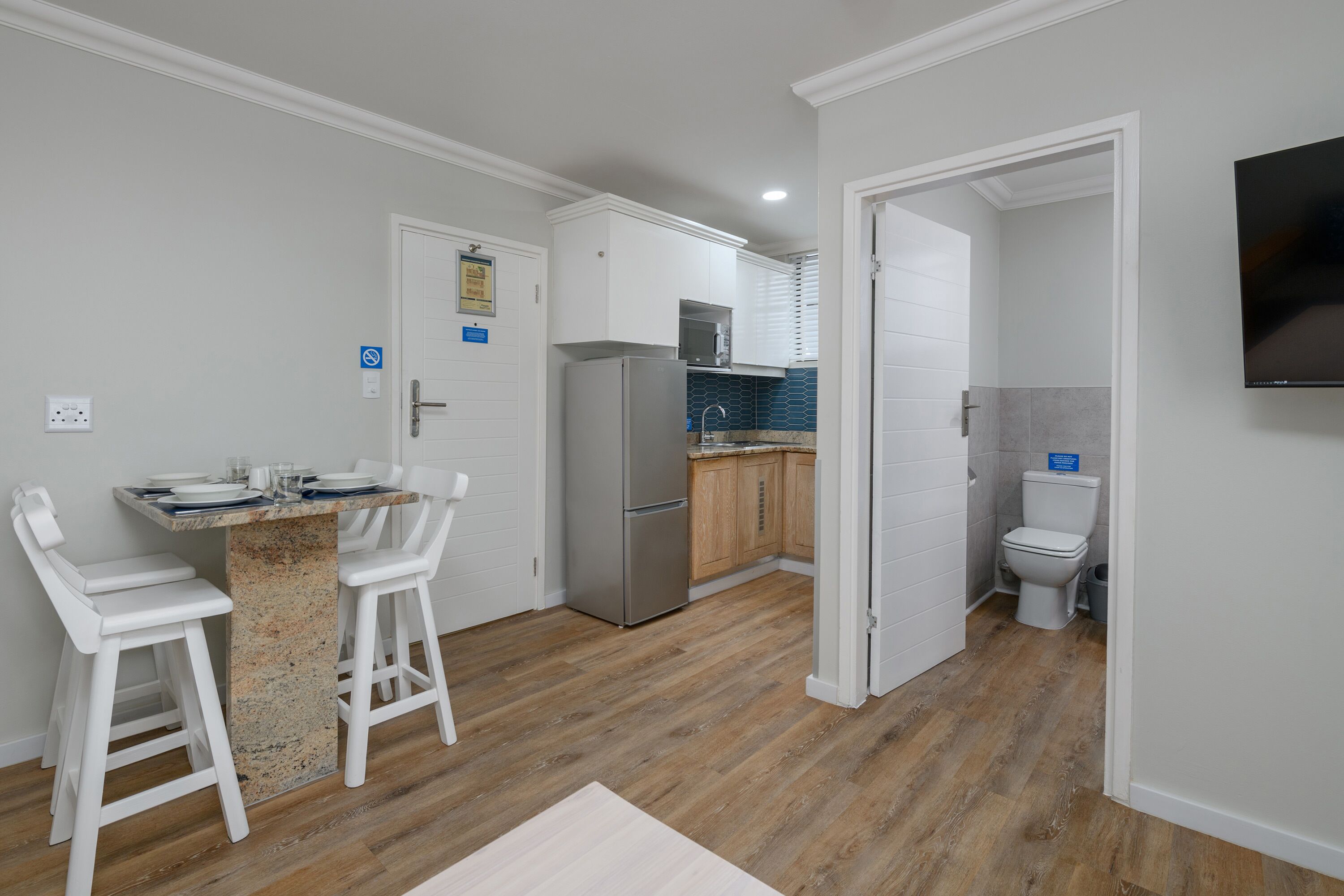 A compact kitchen with white cabinets, a stainless steel refrigerator, and a dining area with white bar stools.