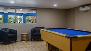 A pool table with two chairs, a small round table, and a window with a view of greenery.