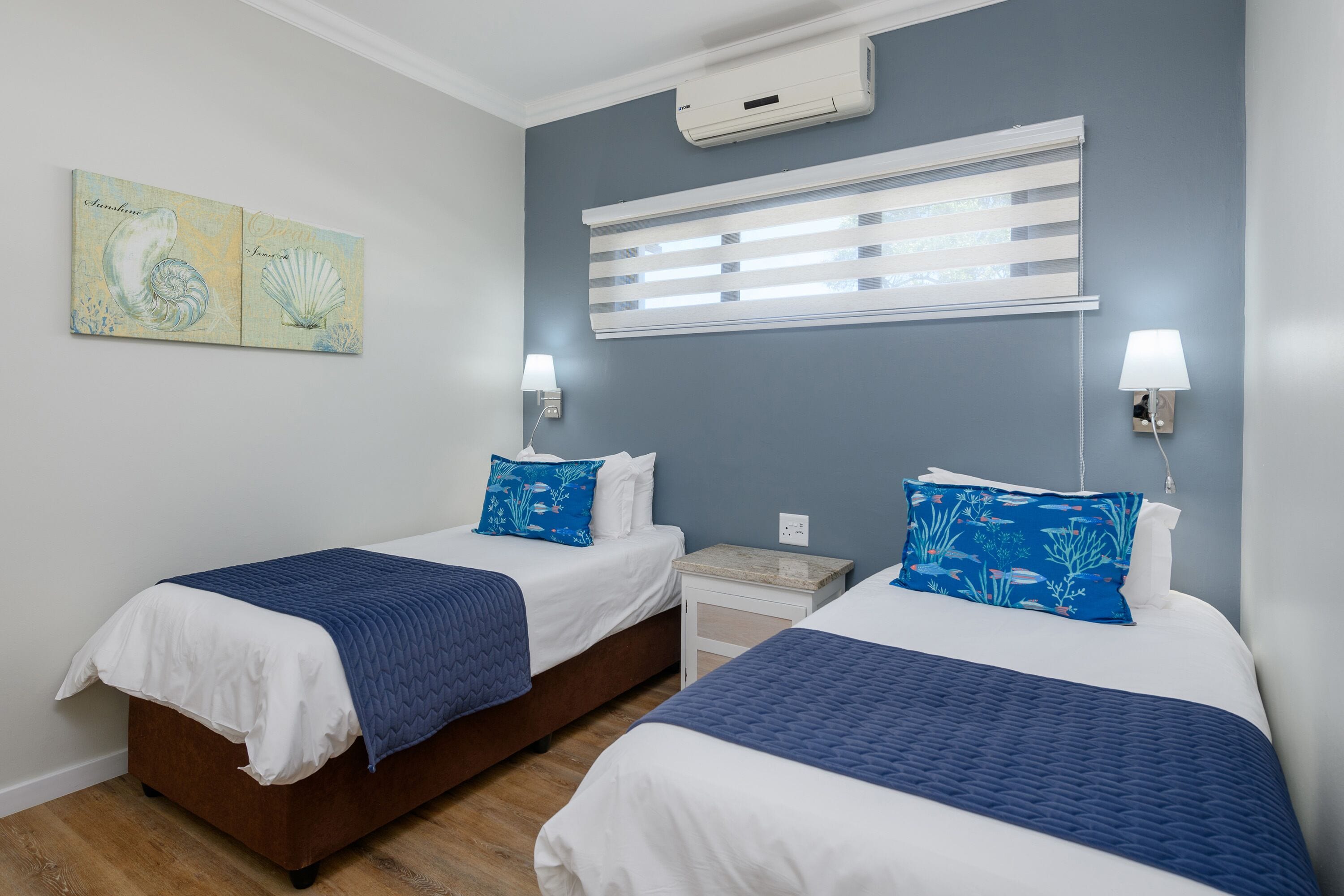 A hotel room with two beds, a nightstand, a wall-mounted air conditioner, and a window with blinds.