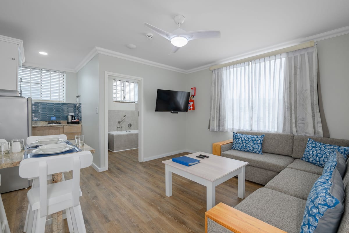 A modern apartment with a dining area, a living room with a sofa and a TV, a kitchen with a window, and a ceiling fan.