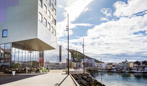 A modern building with a glass facade, a wooden deck, and a marina with boats. - Smarthotel Hammerfest (Hammerfest)