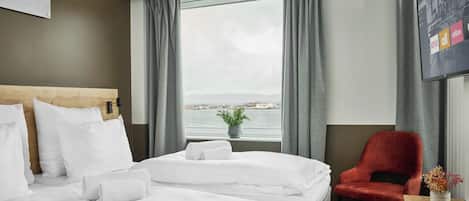 Habitación de hotel con cama, una silla, una mesita con un florero y una ventana con vista al agua.