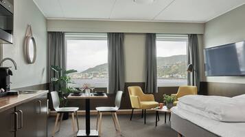A hotel room with a bed, a desk, a chair, a table with a vase and glasses, a potted plant, and a view of a coastal town.