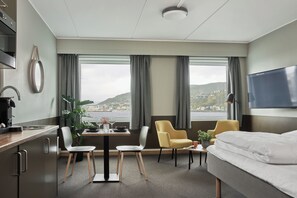 Habitación de hotel con cama, escritorio, silla, mesa con un florero y vasos, una planta en maceta y vistas a un pueblo costero.
