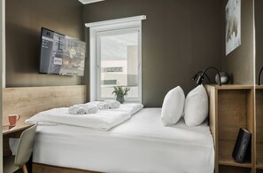 Habitación de hotel con cama, televisor montado en la pared, ventana con vistas a edificios y una mesita con una taza y un libro.