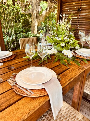 Una mesa de comedor de madera dispuesta con platos blancos, copas de vino y un centro de mesa con flores y follaje blanco.