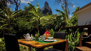 A wooden table with chairs, a pitcher, cups, and a plate of food, set against a backdrop of lush greenery and a rock formation.