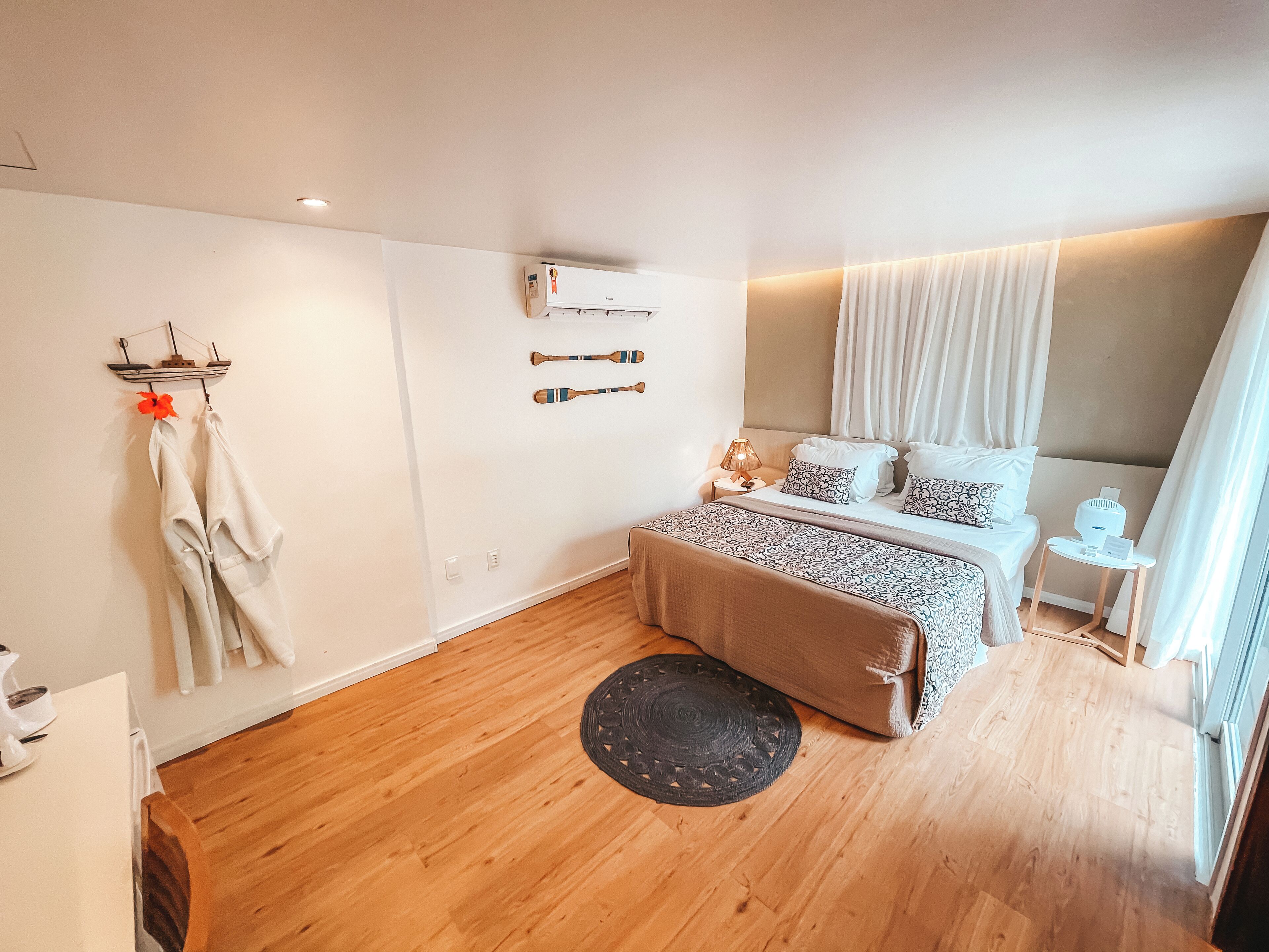 A modern bedroom with a wooden floor, a bed with patterned bedding, a bedside table with a lamp, and a wall-mounted air conditioner.